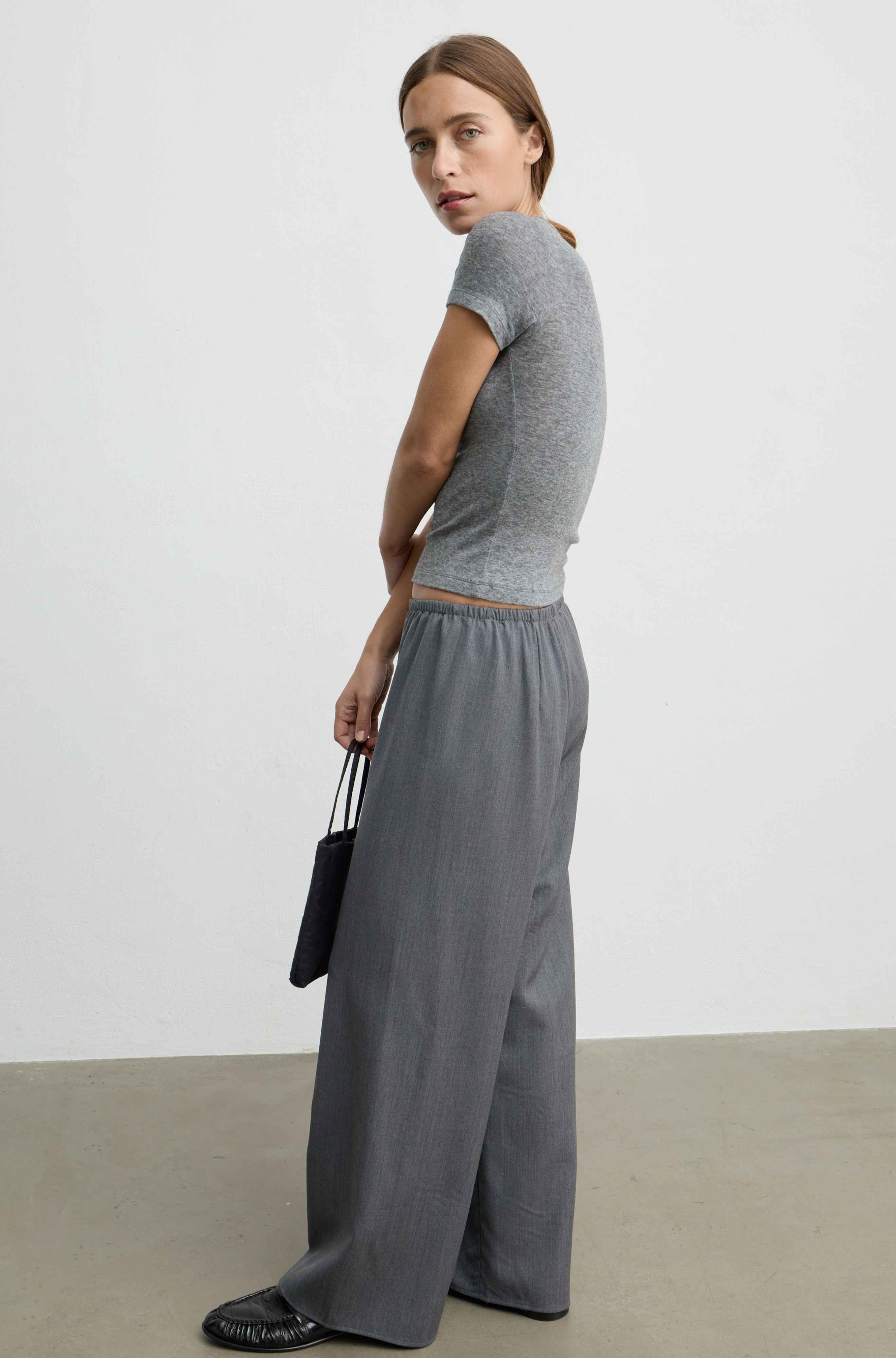 A woman stands in profile wearing a fitted heather gray short-sleeve crop top and matching wide-leg gray trousers, holding a small black handbag against a plain light gray studio background.