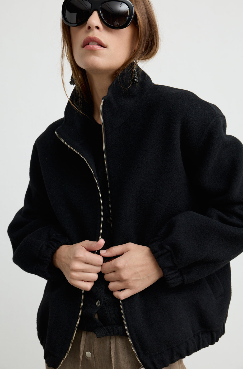 Woman wearing a black wool zip up bomber jacket with brown pants, against a light grey wall