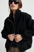 Woman wearing a black wool zip up bomber jacket with brown pants, against a light grey wall