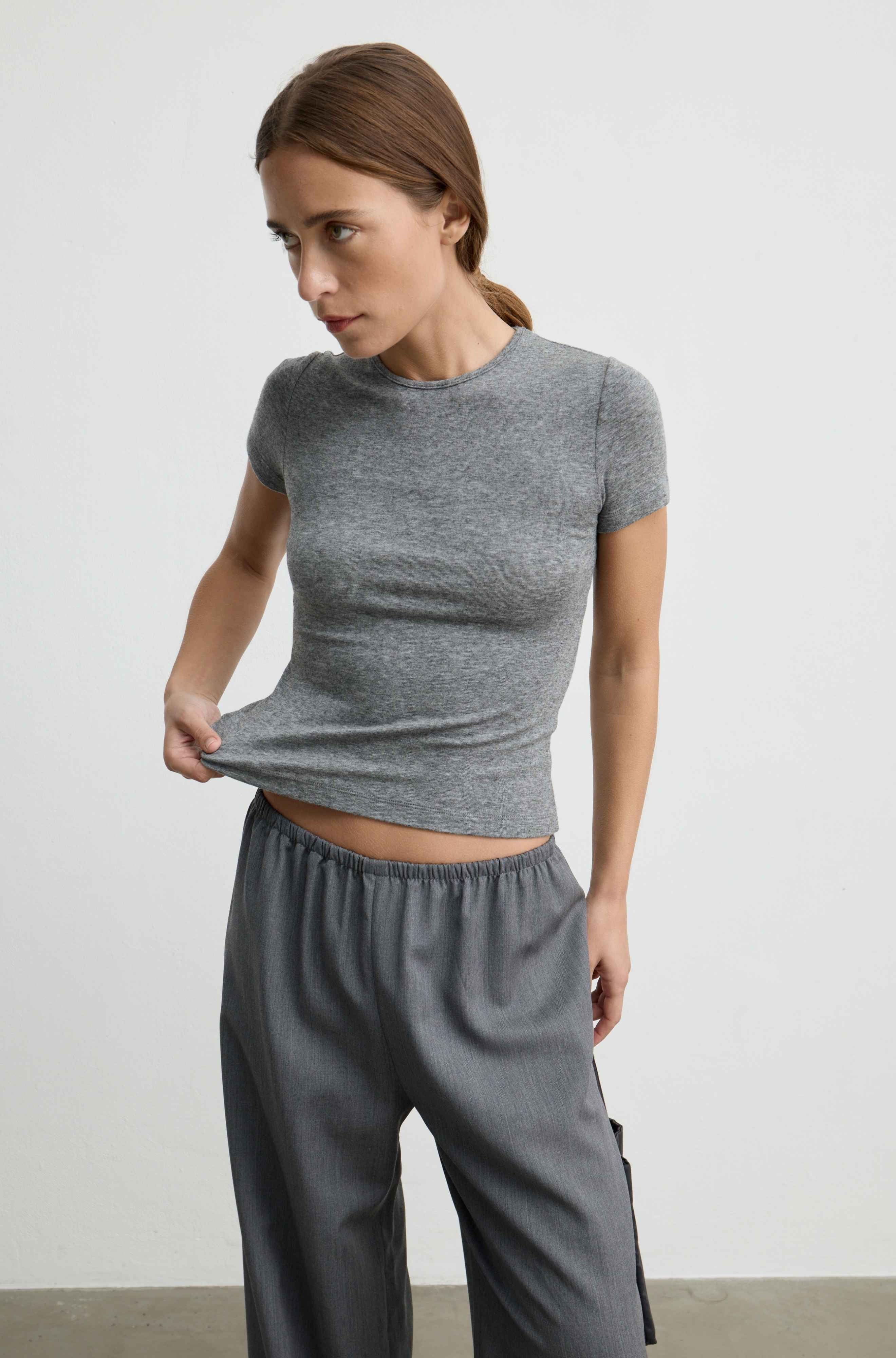 A woman wearing a fitted short-sleeve gray knit top and matching gray pants, standing against a plain light gray wall and pulling the hem of the top slightly outward.