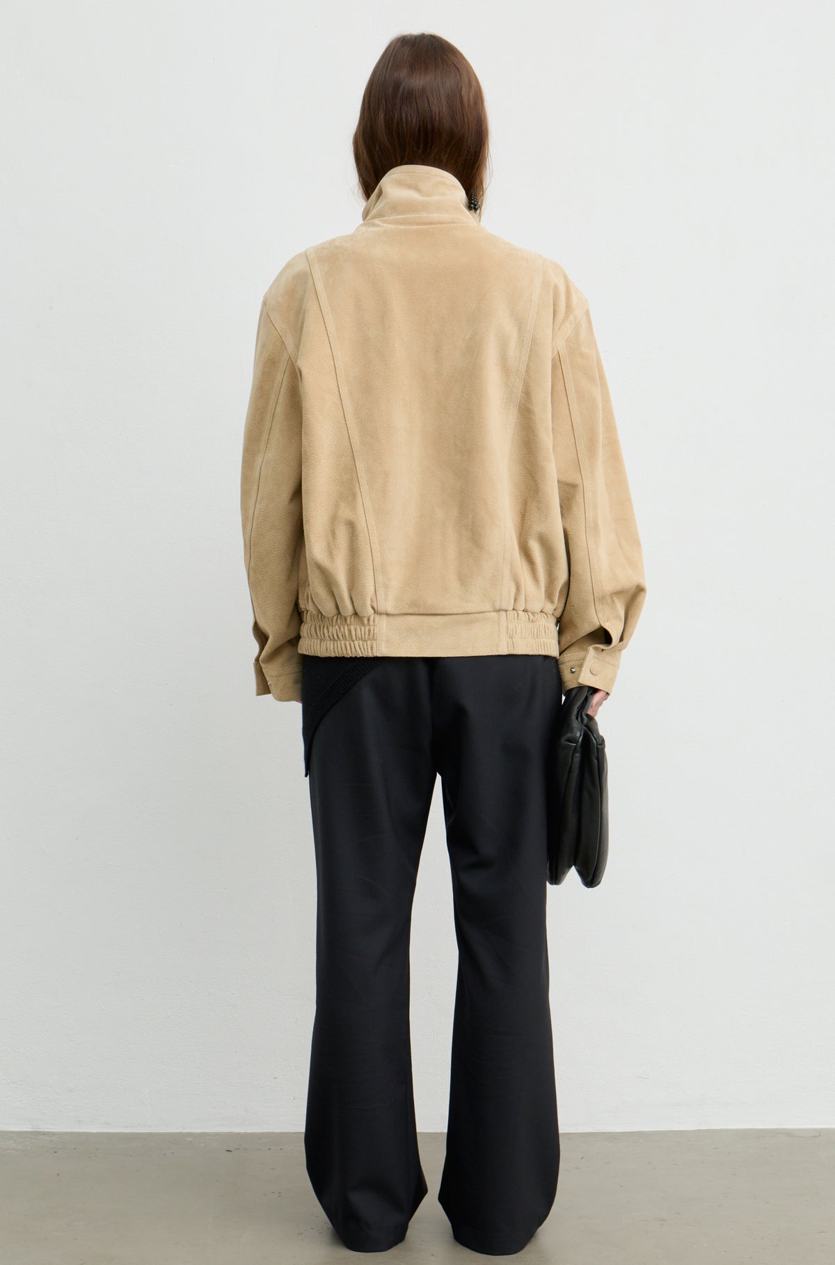 A model shown from the back wearing a beige corduroy jacket and black wide-leg trousers, holding a black clutch bag against a plain white studio background.