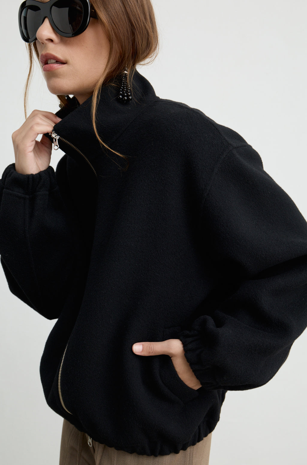 Woman wearing a black wool zip up bomber jacket with brown pants, against a light grey wall