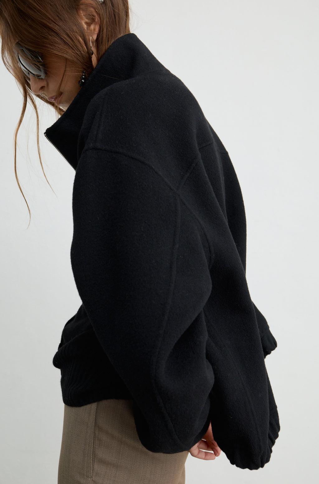 Woman wearing a black wool zip up bomber jacket with brown pants, against a light grey wall