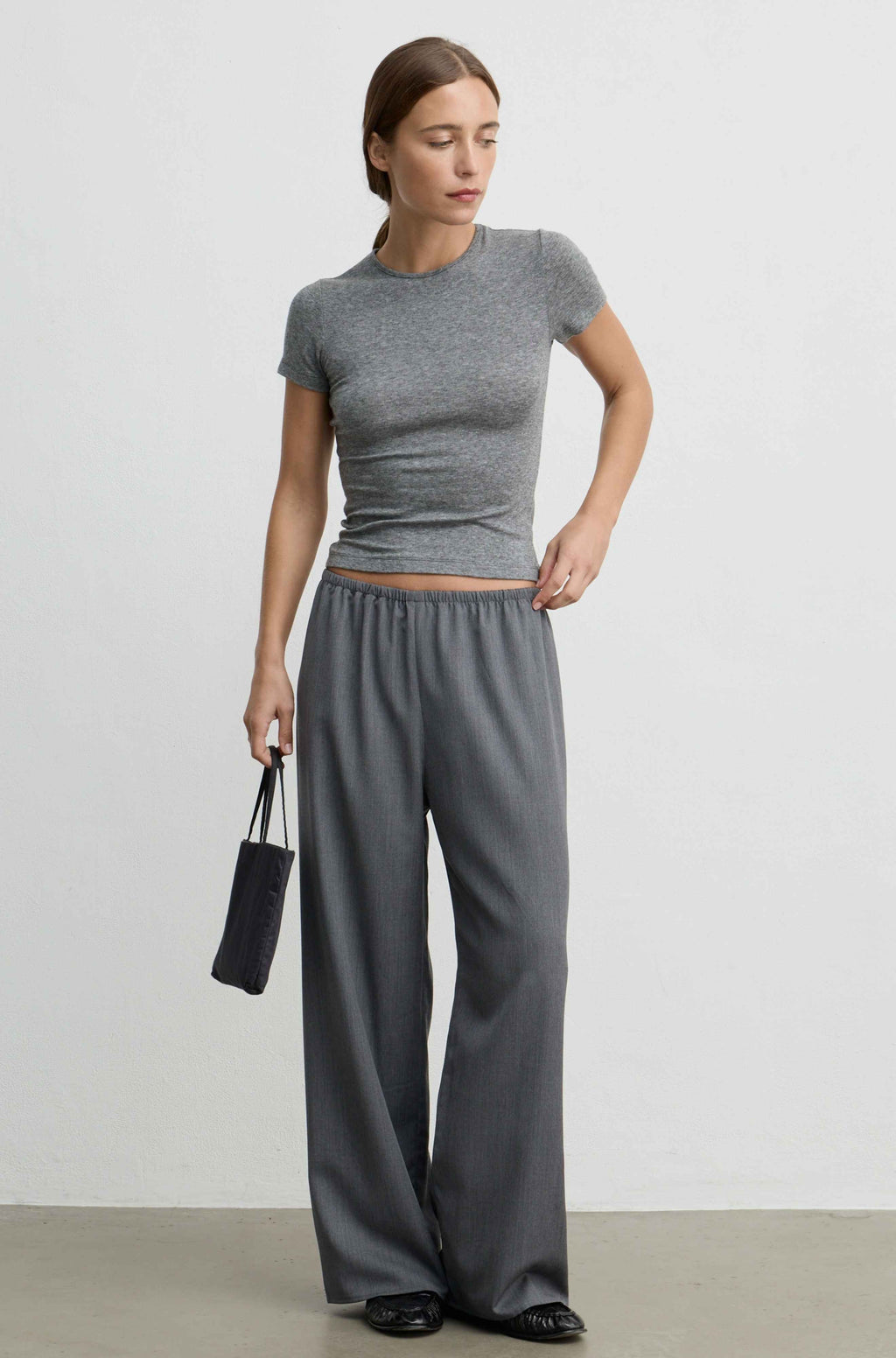 A woman modeling a gray short-sleeve ribbed crop top and matching wide-leg pants, standing against a plain light gray studio wall and holding a small black pouch.