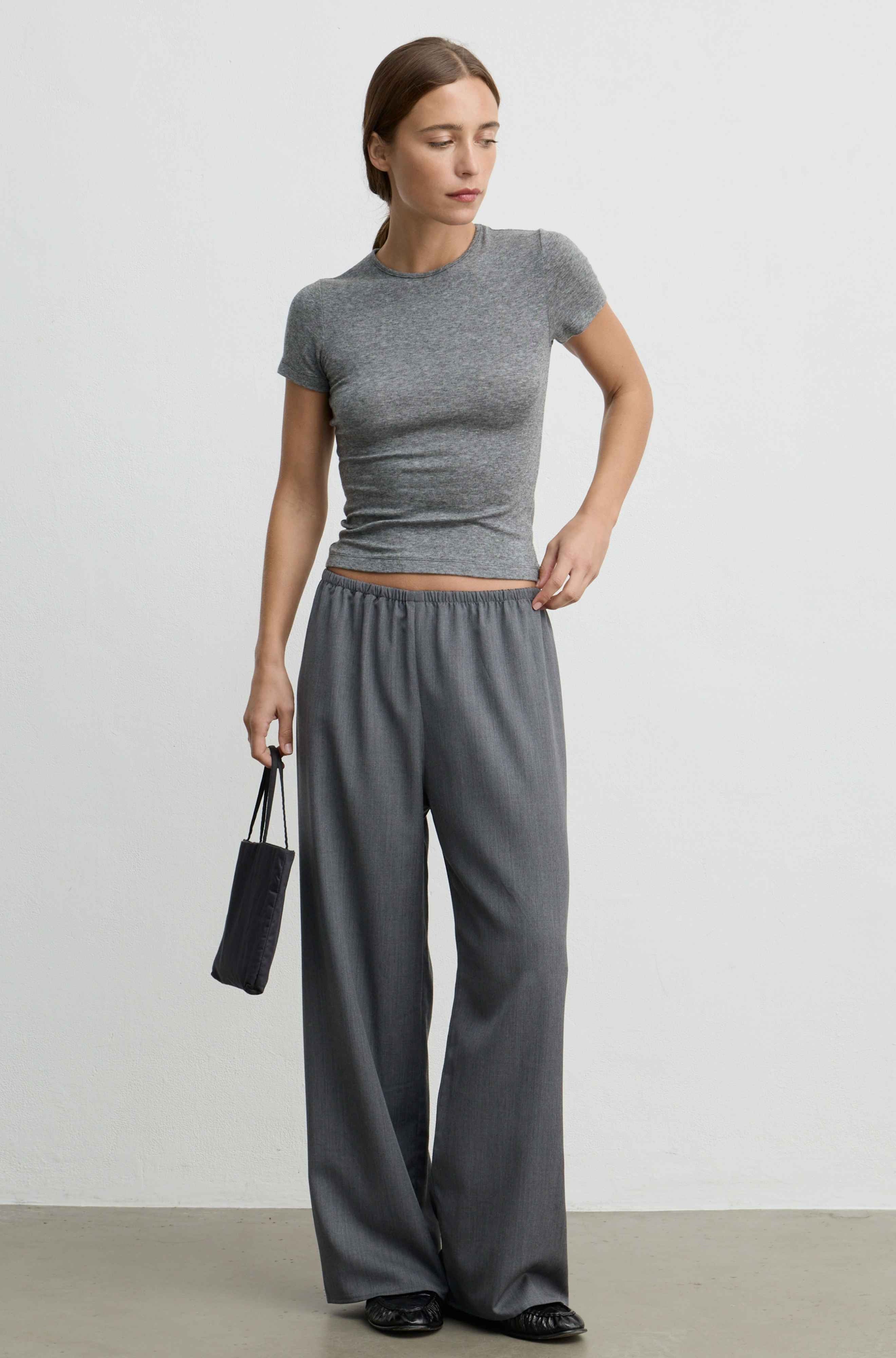 A woman modeling a gray short-sleeve ribbed crop top and matching wide-leg pants, standing against a plain light gray studio wall and holding a small black pouch.