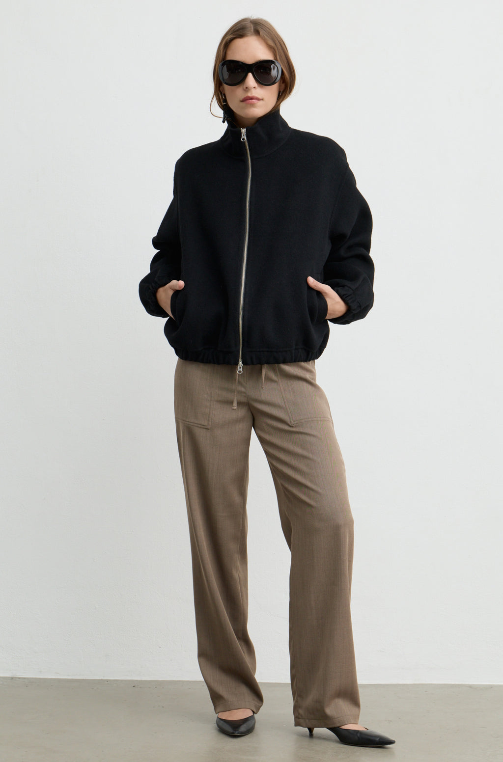 Woman wearing a black wool zip up bomber jacket with brown pants, against a light grey wall