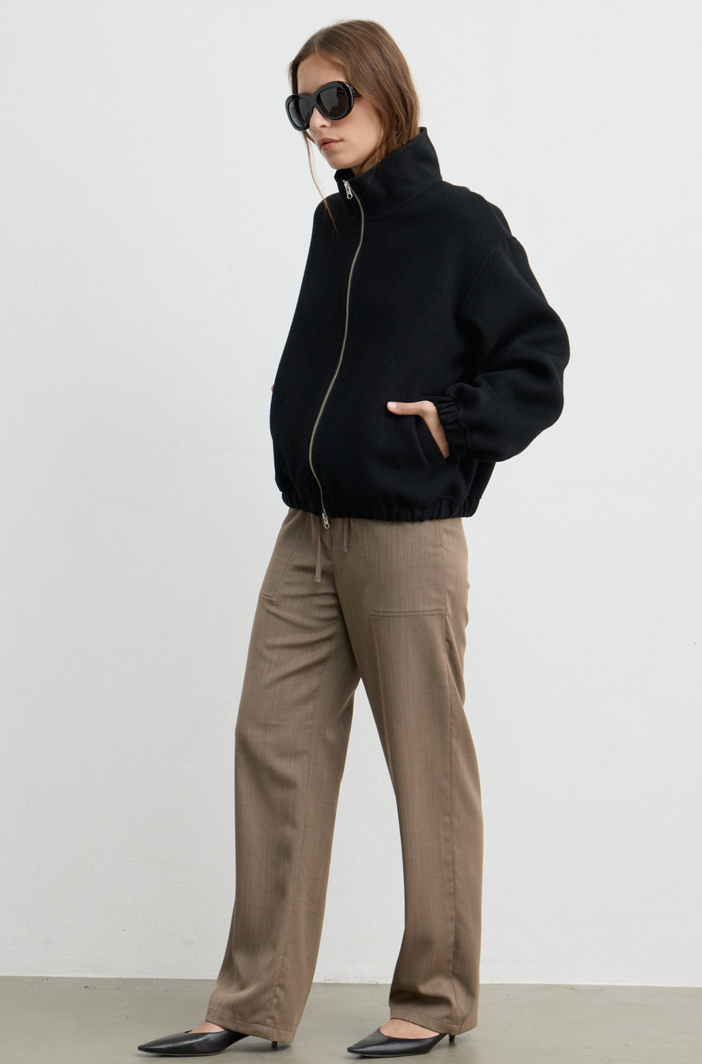Woman wearing a black wool zip up bomber jacket with brown pants, against a light grey wall