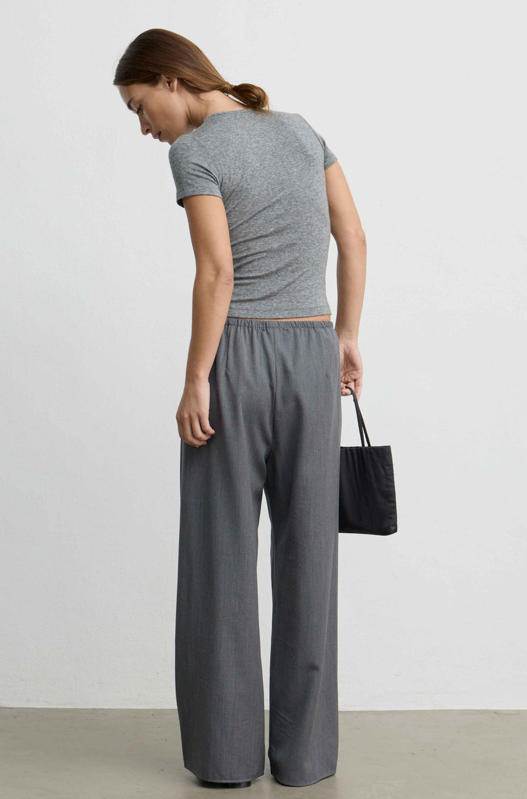 A woman wearing a gray short-sleeve cropped top and matching wide-leg trousers, shown from the back while holding a black tote bag against a plain light gray wall.