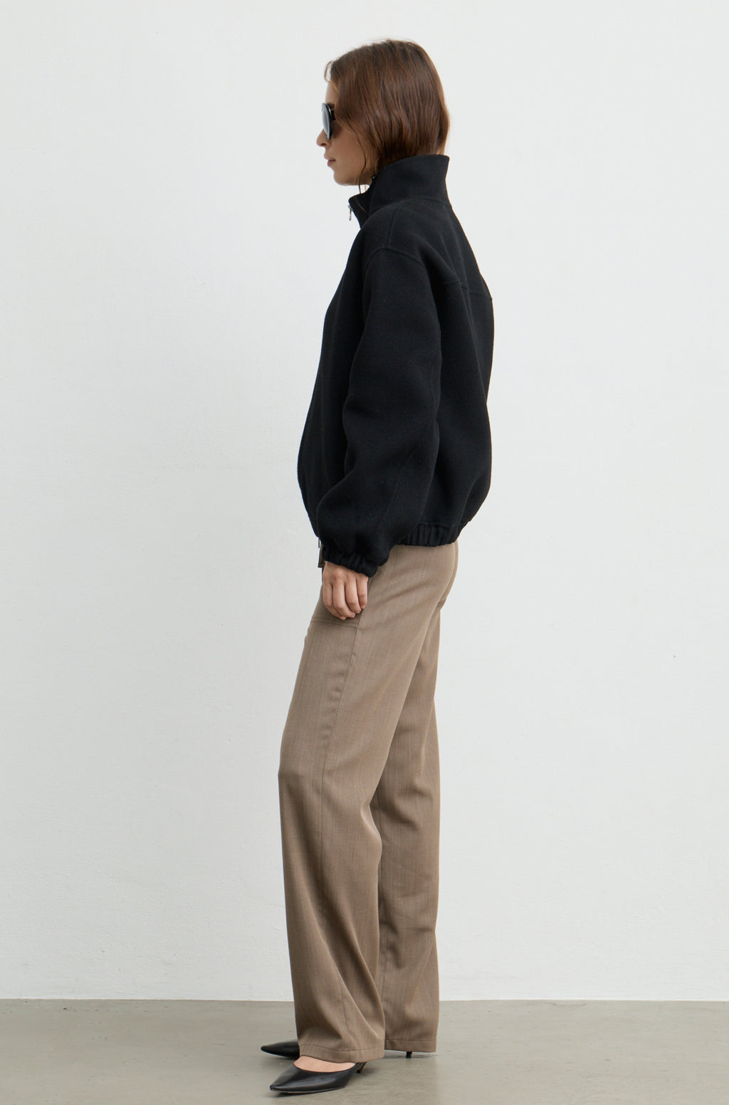 Woman wearing a black wool zip up bomber jacket with brown pants, against a light grey wall