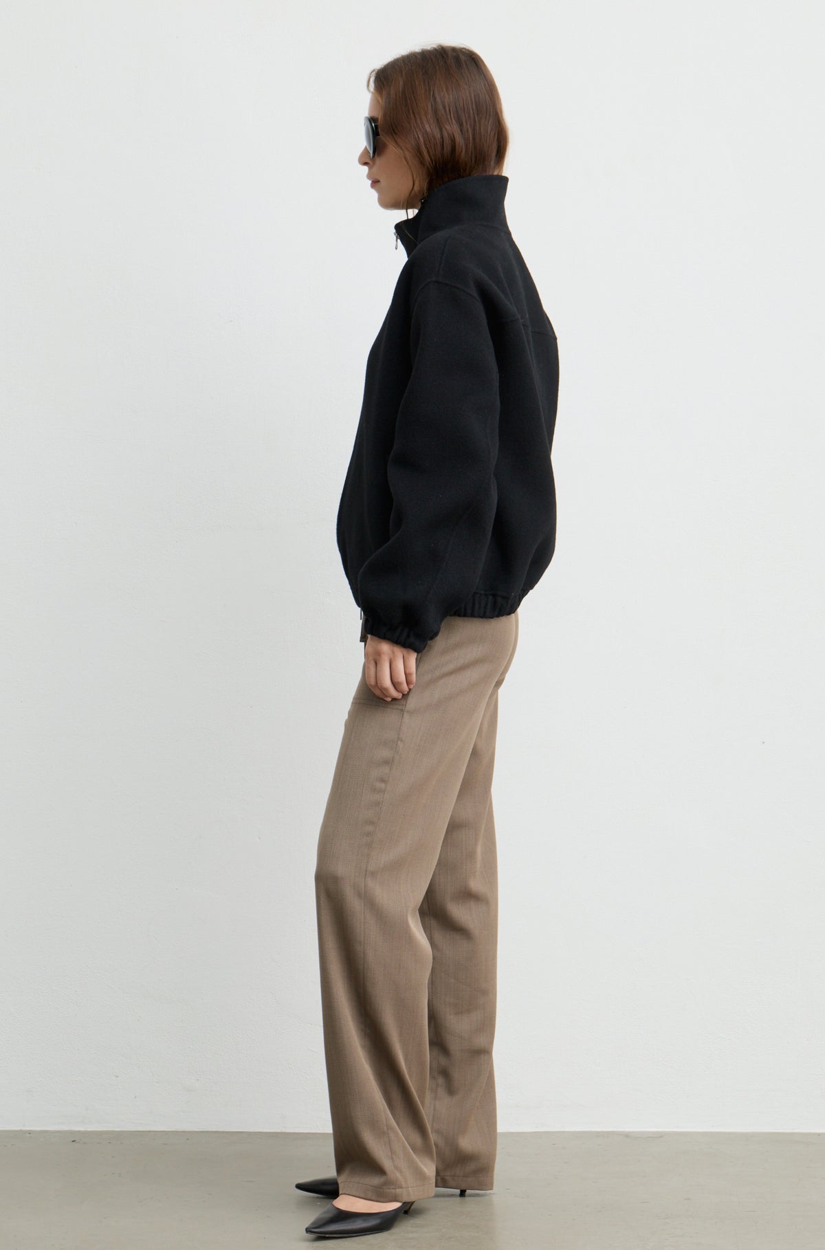 Woman wearing a black wool zip up bomber jacket with brown pants, against a light grey wall