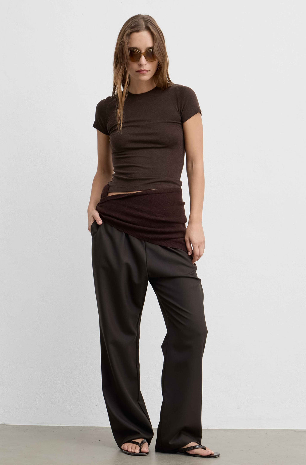 A woman modeling a dark brown short-sleeve top and matching dark brown draped pants against a plain light gray studio background.