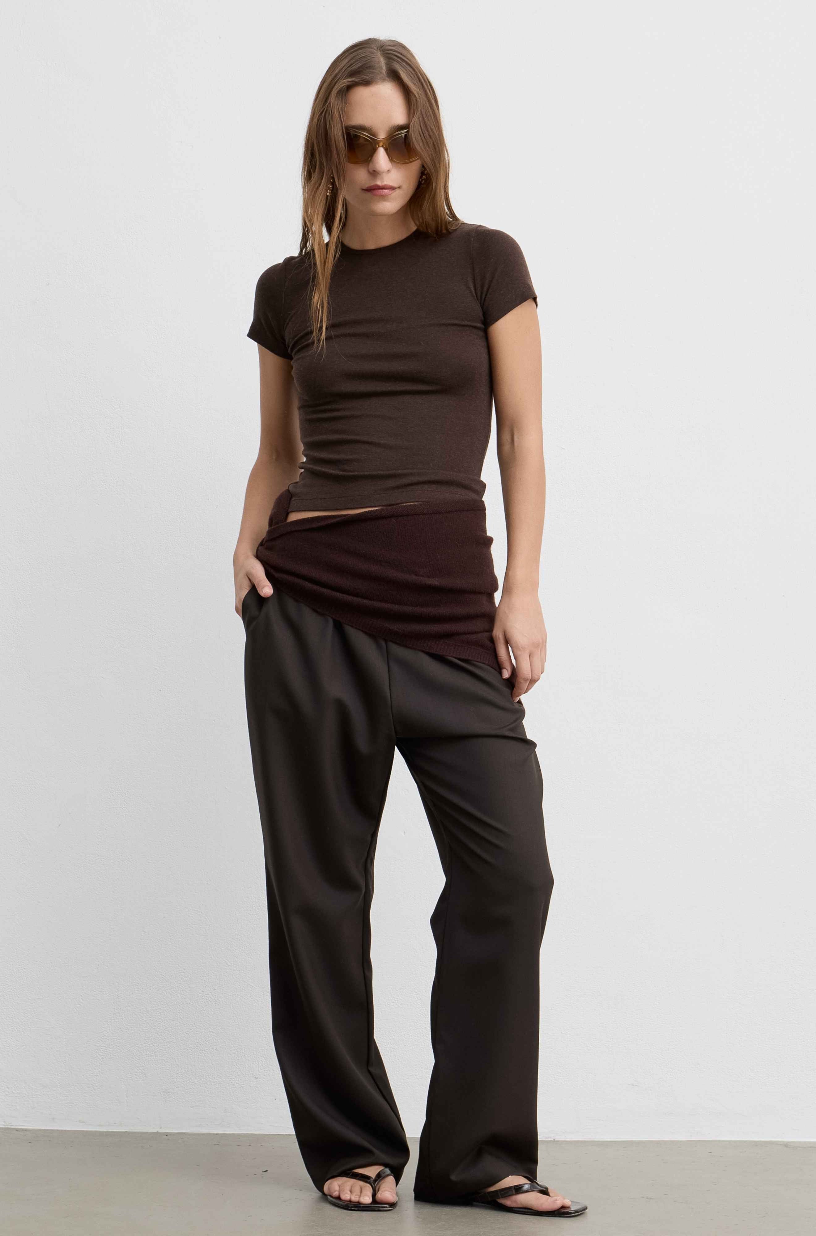 A woman modeling a dark brown short-sleeve top and matching dark brown draped pants against a plain light gray studio background.