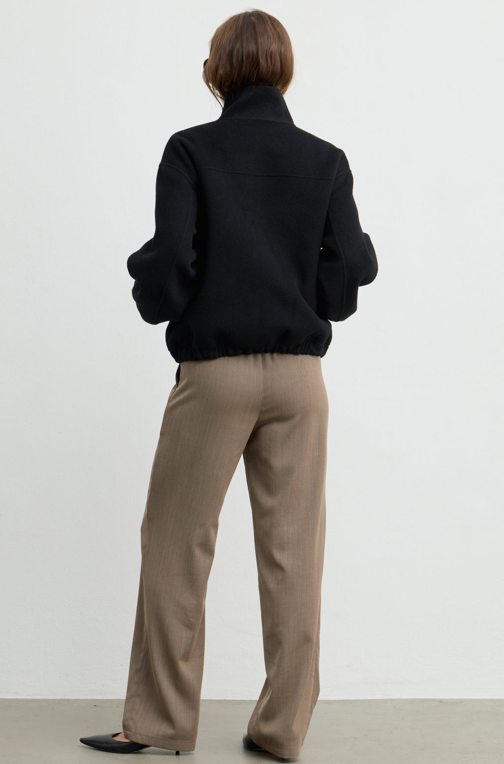 Woman wearing a black wool zip up bomber jacket with brown pants, against a light grey wall