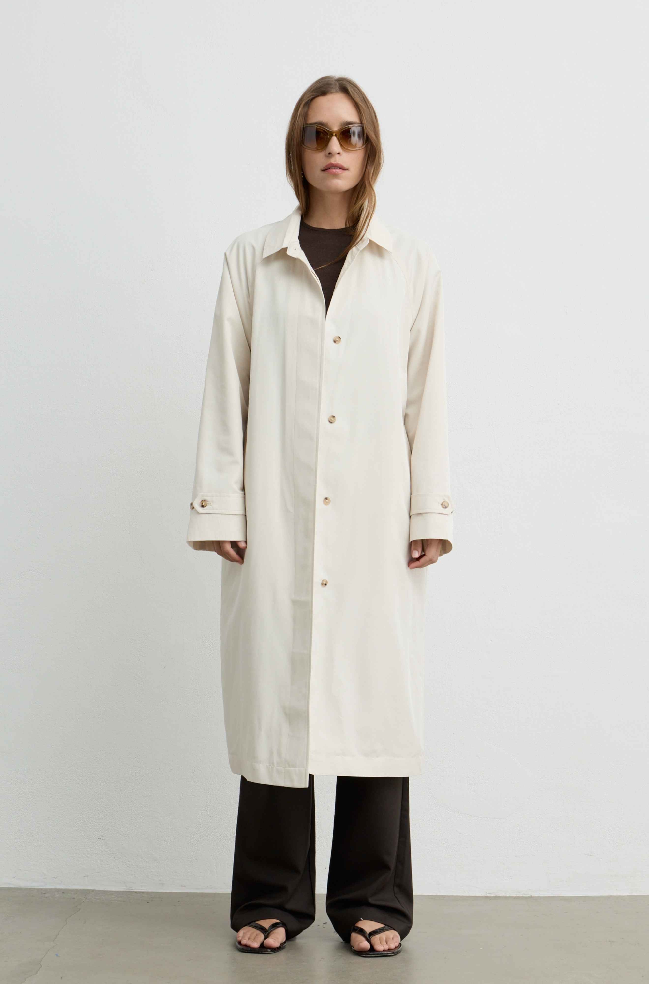 A woman modeling a long cream-colored shirt jacket with gold buttons, worn over a black top and black wide-leg pants, standing against a plain white studio background.