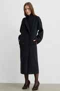 Woman wearing a dark grey button up coat with brown heels against a light grey wall
