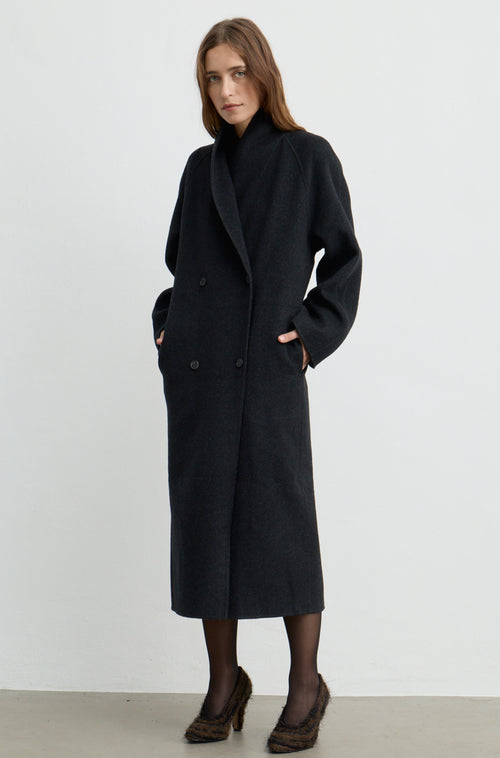 Woman wearing a dark grey button up coat with brown heels against a light grey wall