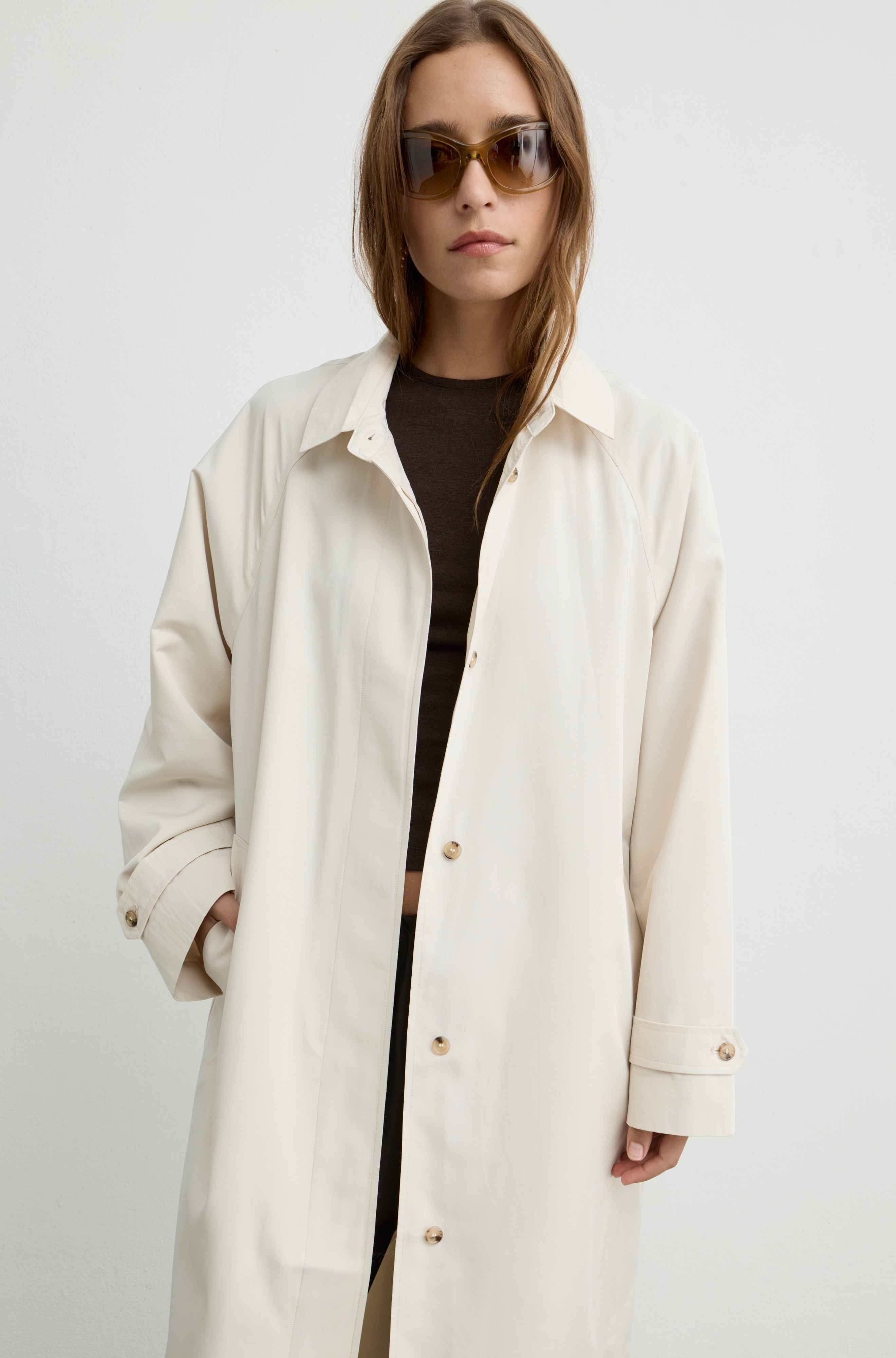 A woman wearing an oversized cream-colored coat over a dark brown top, styled with sunglasses, against a plain light gray background.