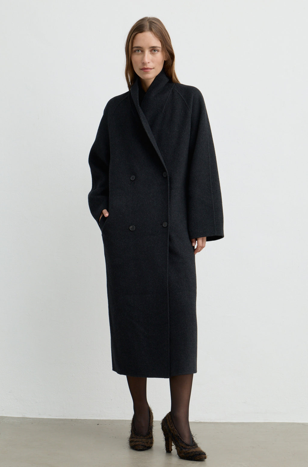 Woman wearing a dark grey button up coat with brown heels against a light grey wall