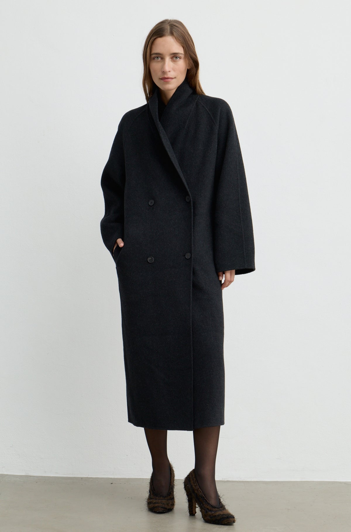 Woman wearing a dark grey button up coat with brown heels against a light grey wall