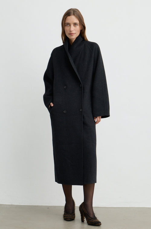 Woman wearing a dark grey button up coat with brown heels against a light grey wall