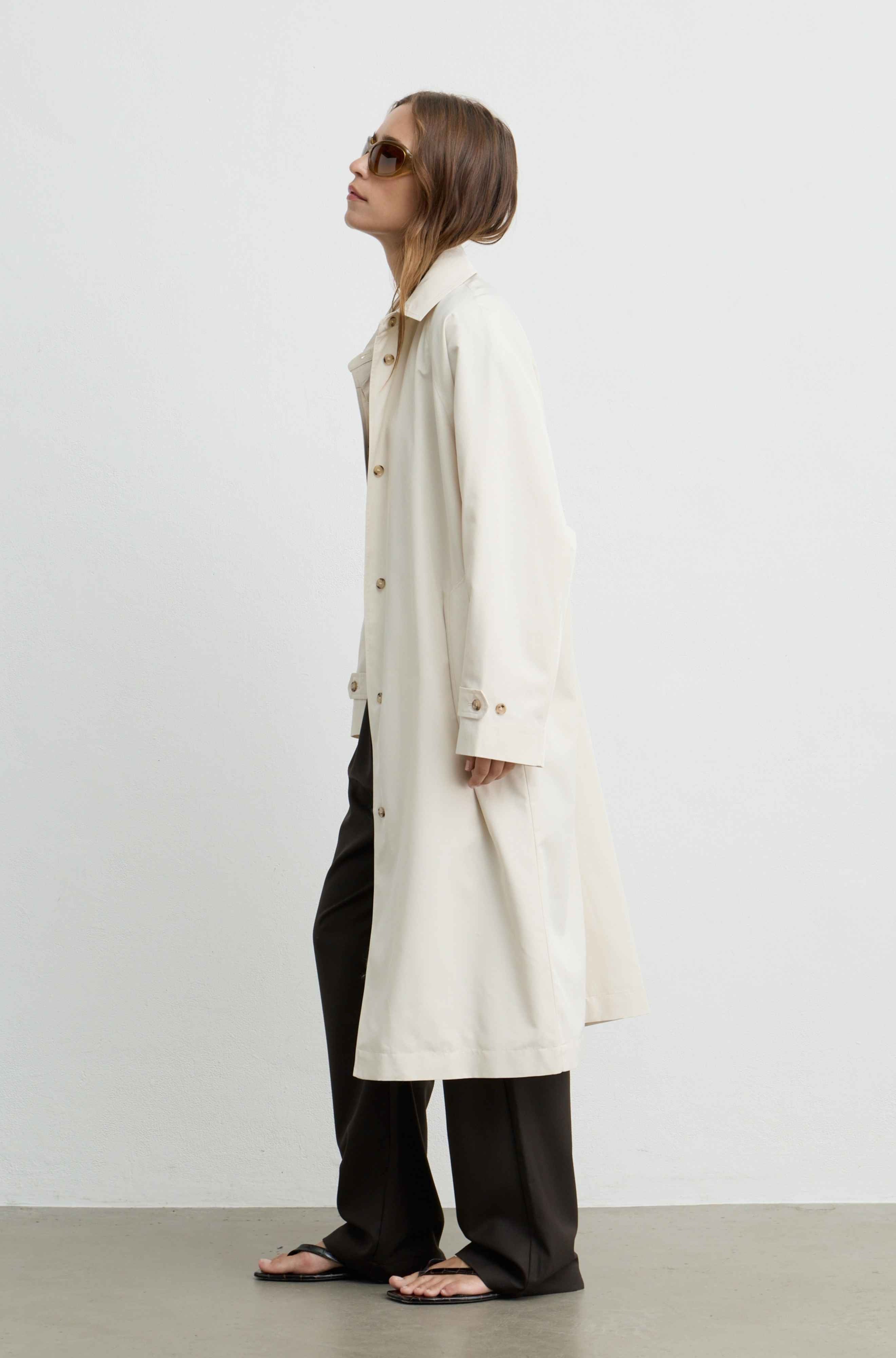 A woman wearing a long cream trench coat, loose black pants, and sandals, standing in profile against a plain light gray studio background.