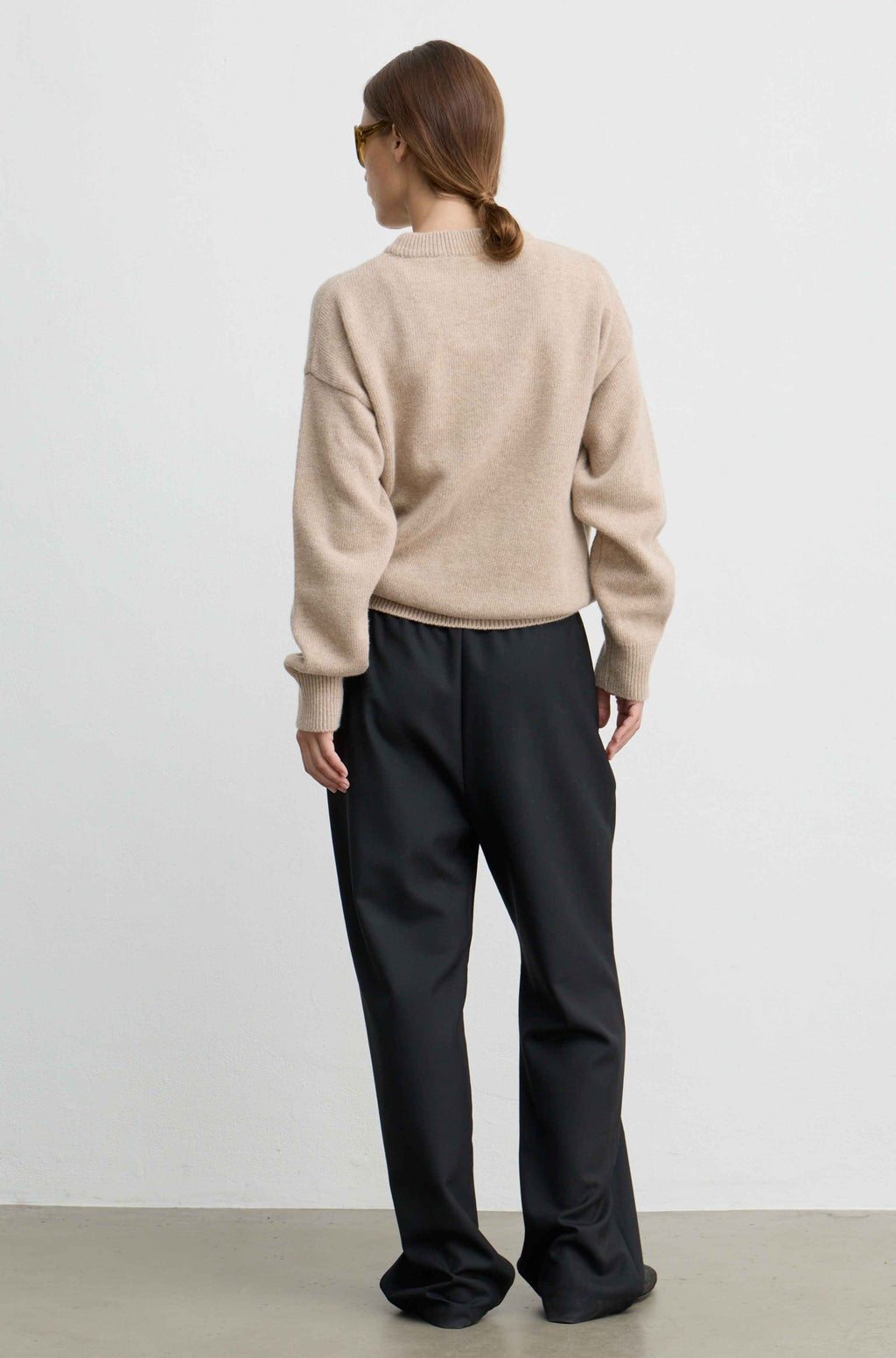 A woman shown from the back wearing a beige knit sweater and black wide-leg trousers, standing against a plain light gray studio wall.