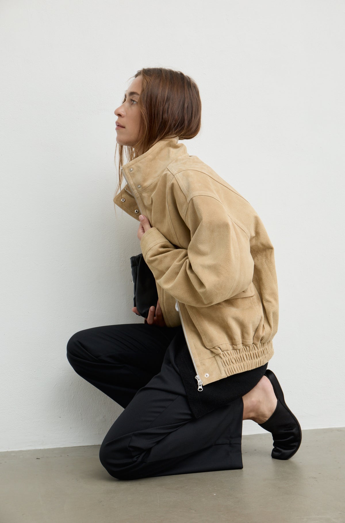 A woman kneels on a concrete floor against a white wall, wearing a tan corduroy jacket, black pants, and black socks.