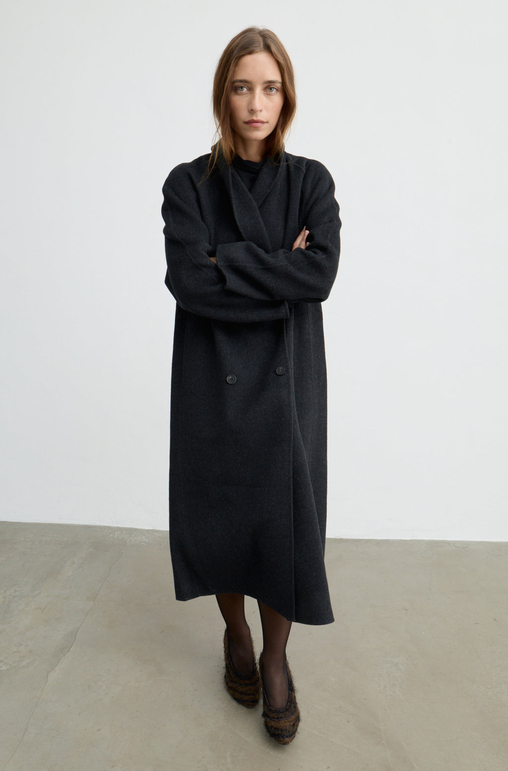 Woman wearing a dark grey button up coat with brown heels against a light grey wall