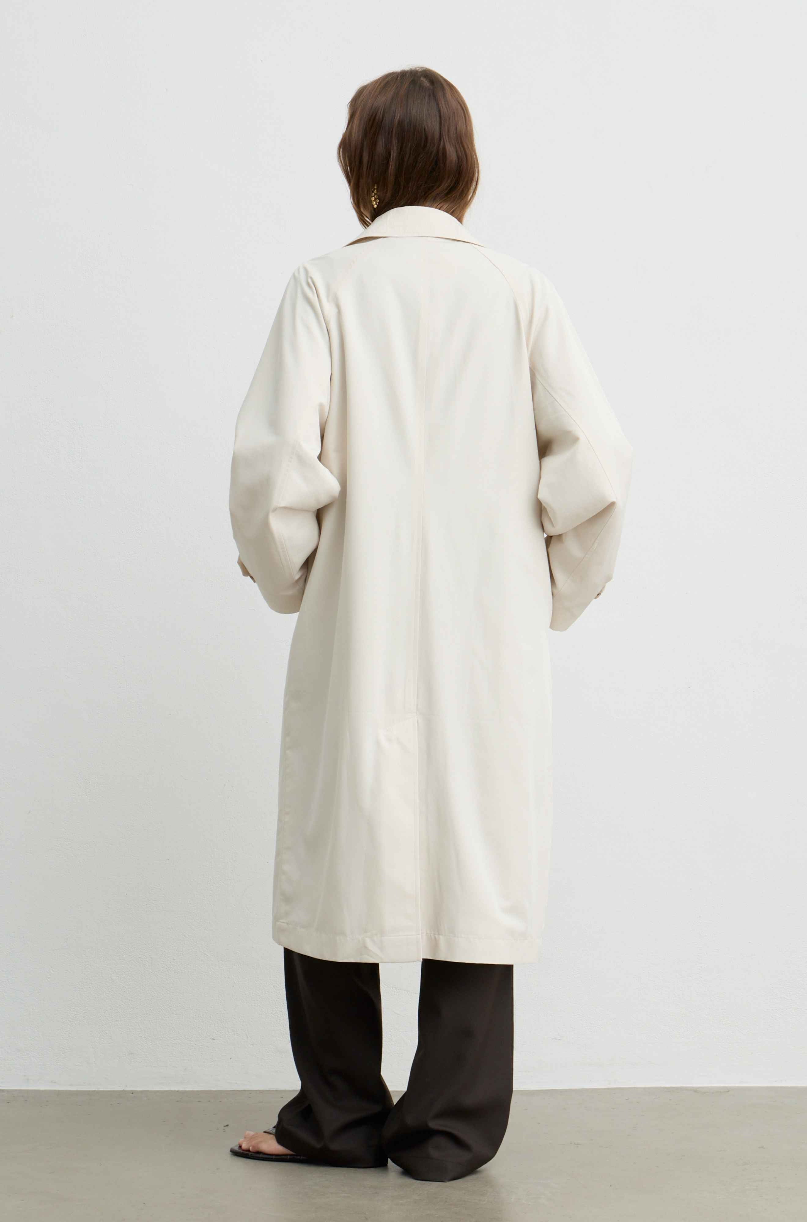 A model viewed from behind wearing a long, oversized cream-colored coat over dark wide-leg trousers, standing against a plain light gray wall.