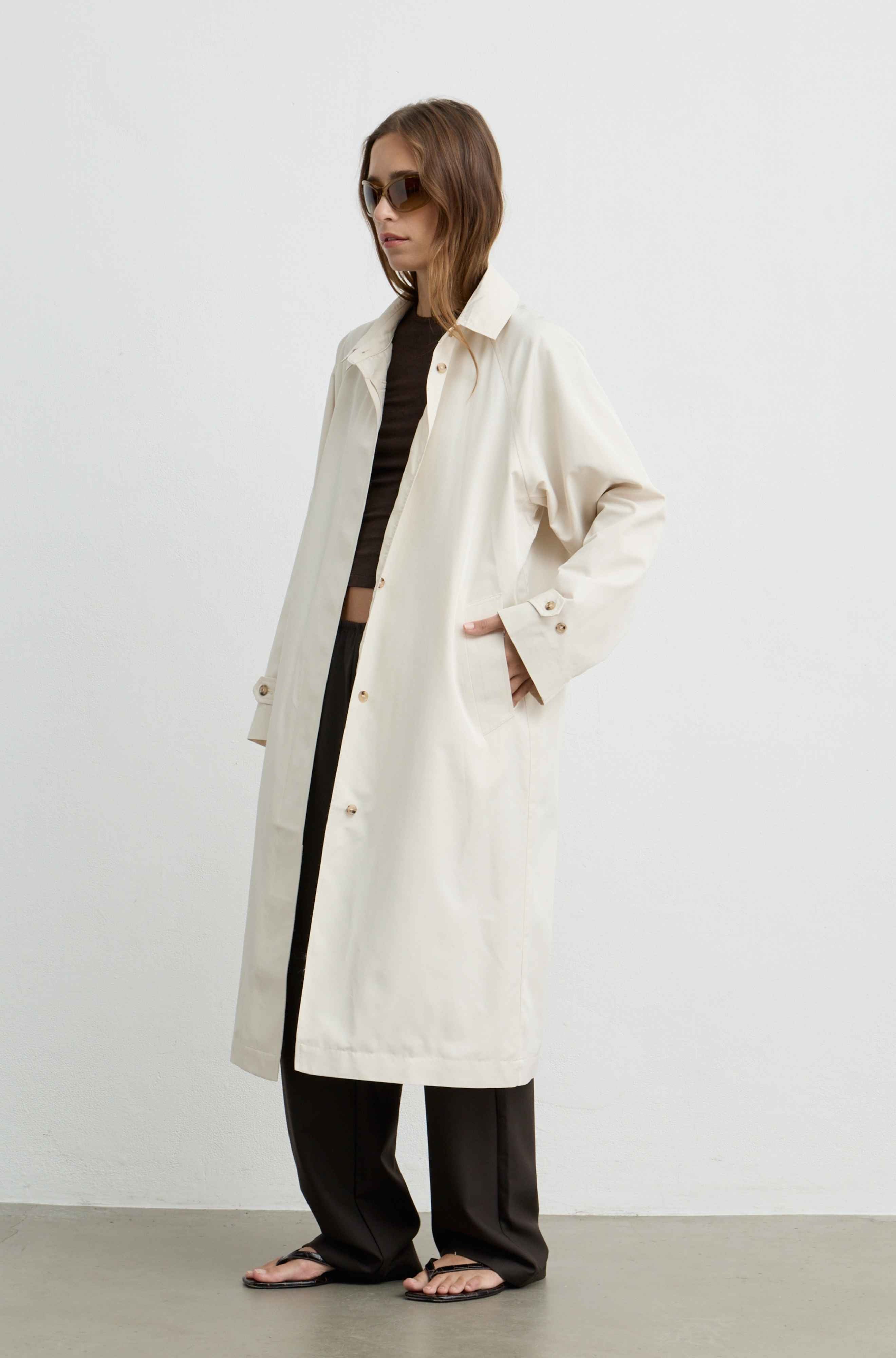 A woman models a long, oversized off-white coat over a black crop top and black wide-leg pants, standing against a plain light gray studio background.