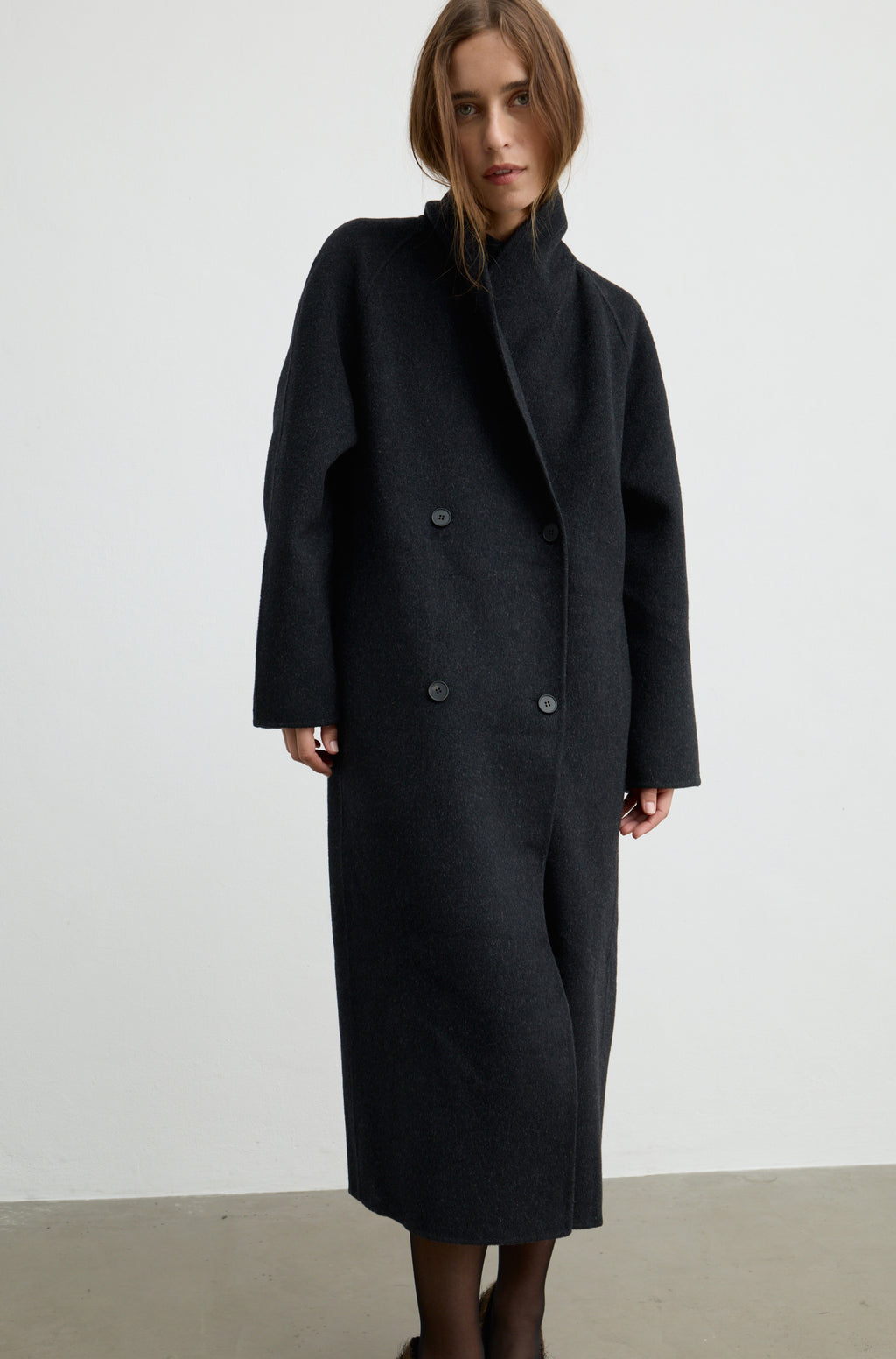 Woman wearing a dark grey button up coat with brown heels against a light grey wall