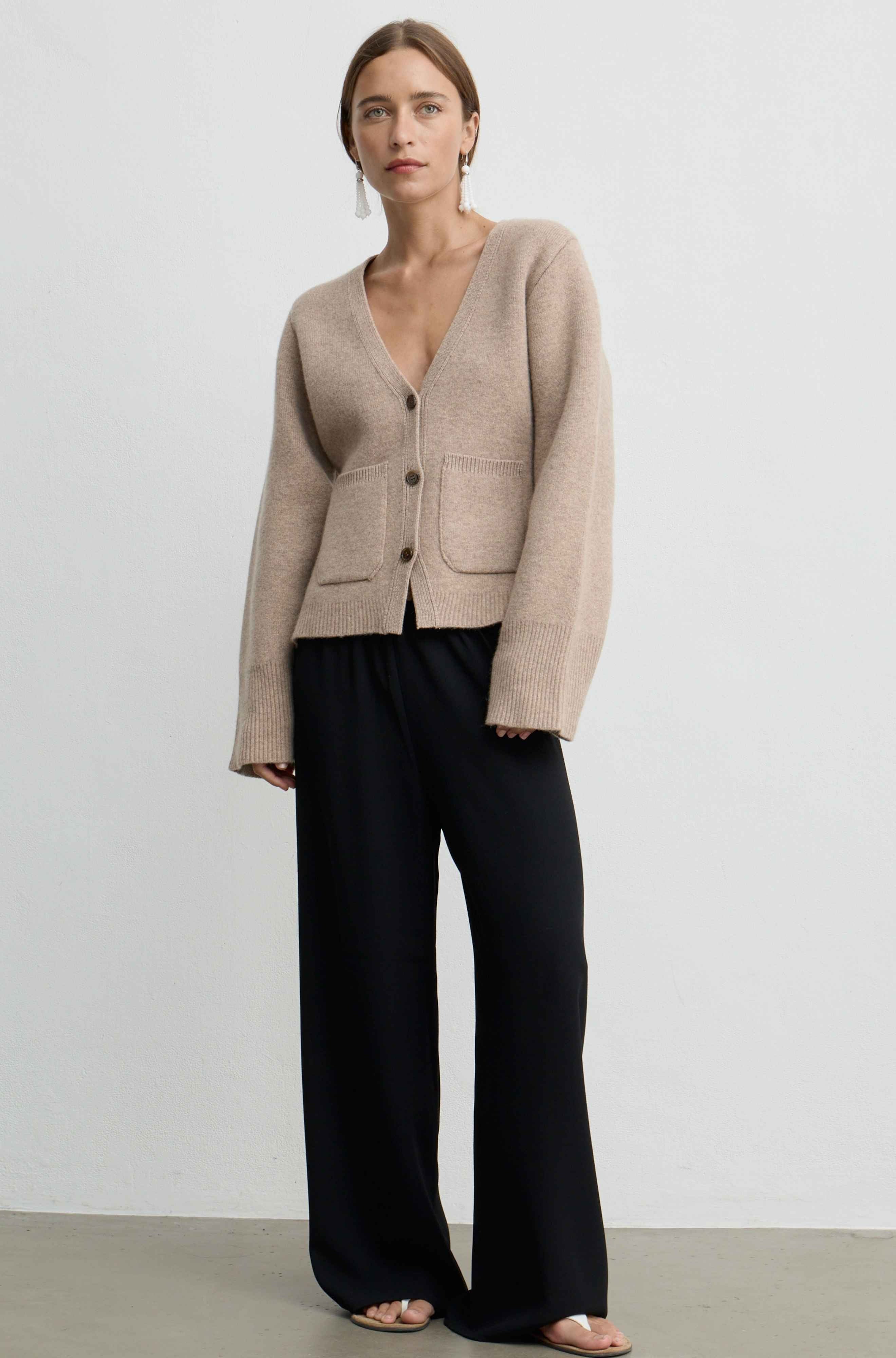 A woman modeling a light beige knit cardigan with a V-neck and front buttons, paired with black wide-leg trousers, against a plain light gray studio background.