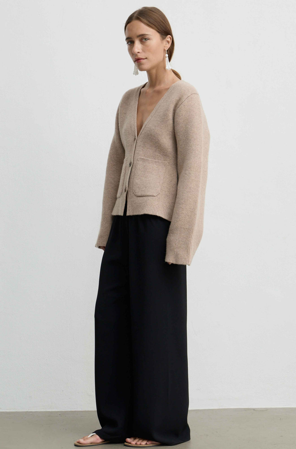 A woman modeling a light beige knit cardigan with a deep V-neck and front pockets, paired with wide-leg black trousers and flat sandals, set against a plain light gray studio background.