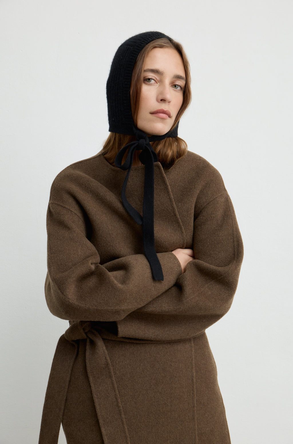 Woman wearing a wool knit hat tied across the neck, with a brown wool coat against a light grey wall