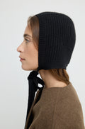 Woman wearing a wool knit hat tied across the neck, with a brown wool coat against a light grey wall