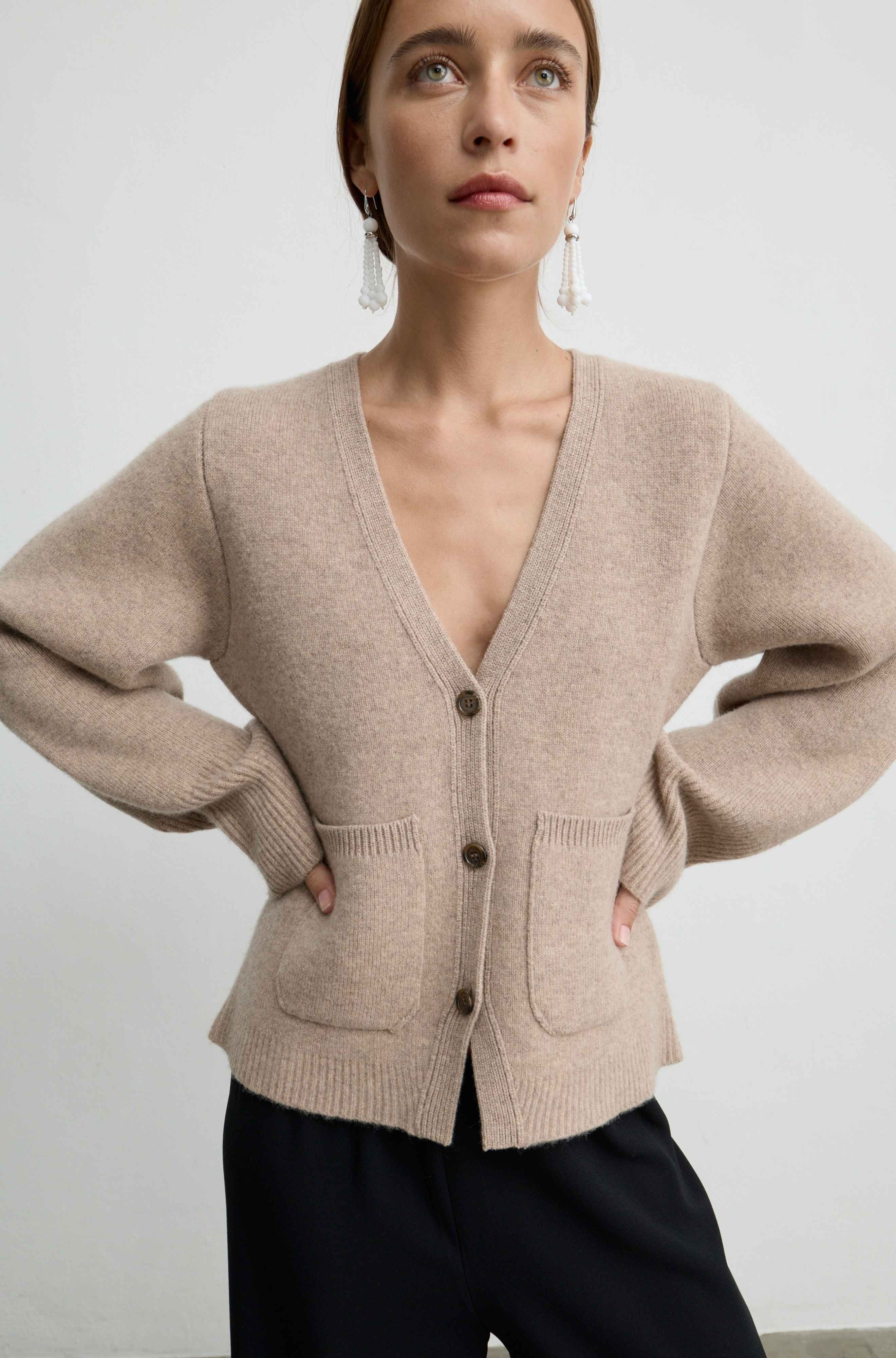 A woman wearing a light beige knit cardigan with a deep V-neck, front buttons, and patch pockets, paired with dark pants against a plain light gray background.