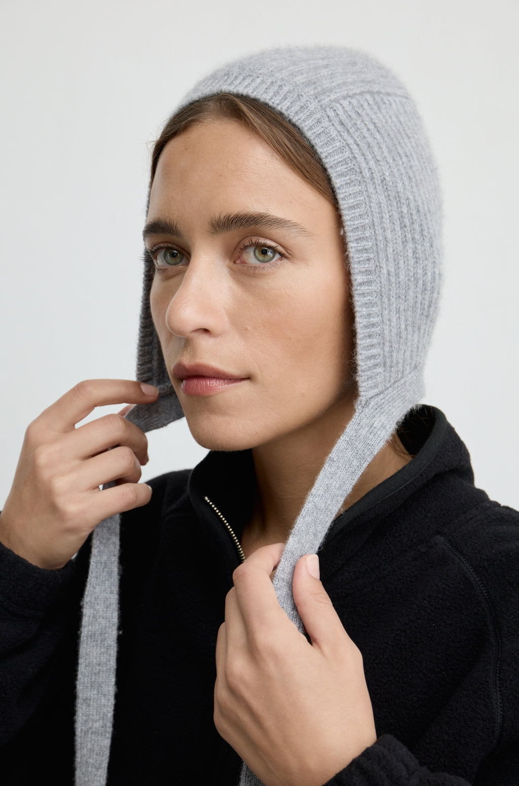 Woman wearing a grey knit hat with ties around the neck with a black flannel sweatshirt against a light grey wall
