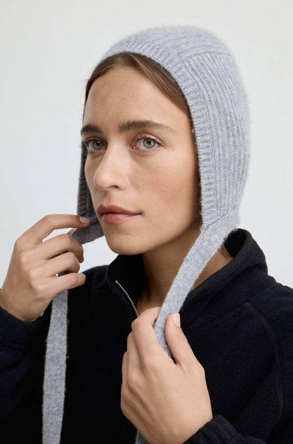 Woman wearing a grey knit hat with ties around the neck with a black flannel sweatshirt against a light grey wall