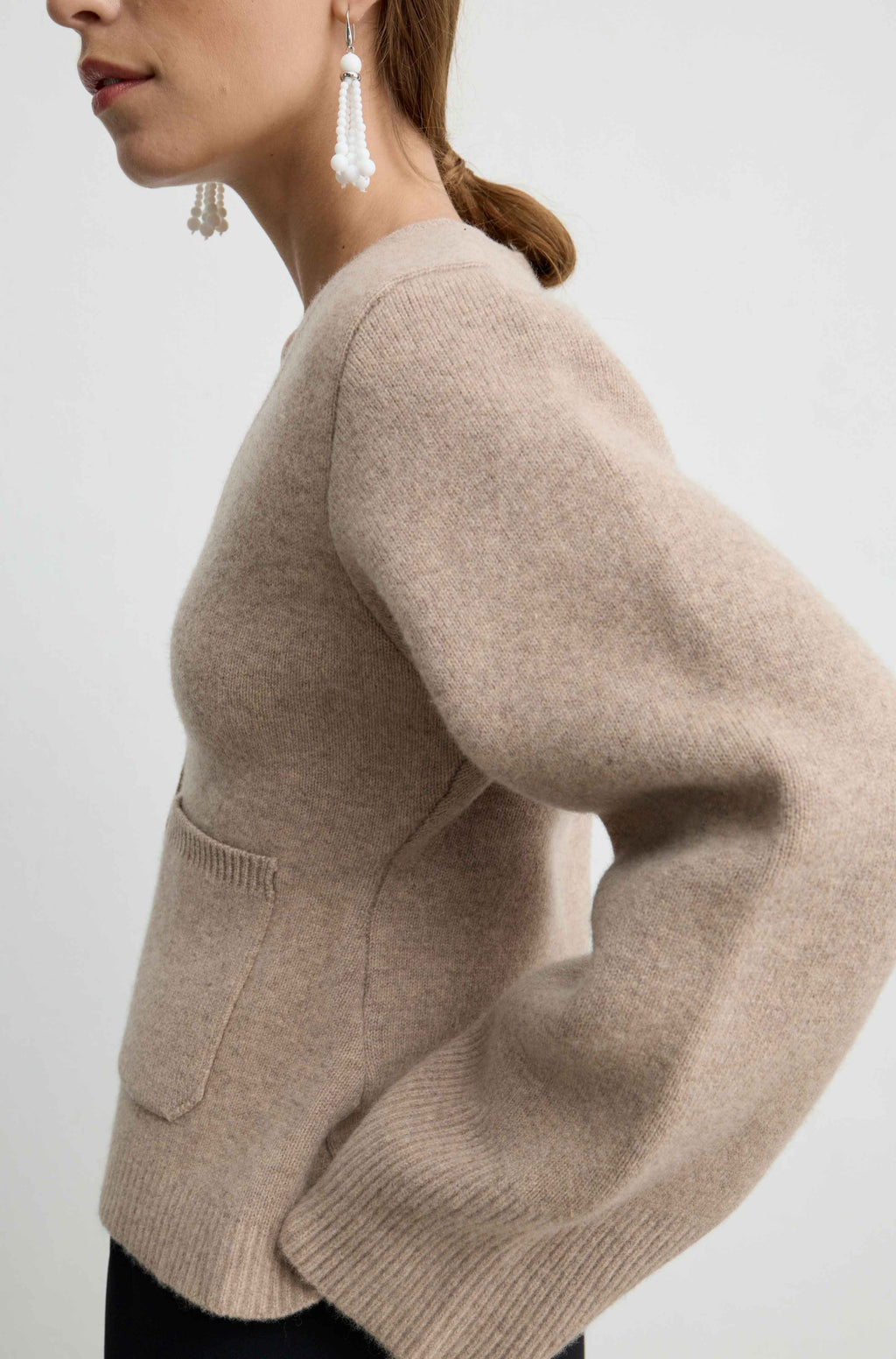 A close-up side view of a woman wearing a light beige knit sweater with a front patch pocket, paired with white tassel earrings, against a plain light gray background.