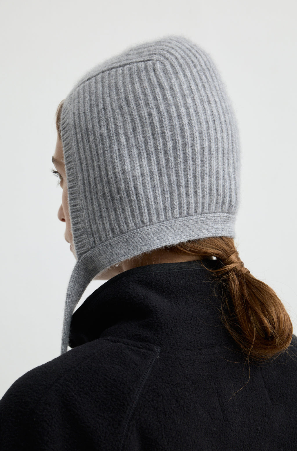 Woman wearing a grey knit hat with ties around the neck with a black flannel sweatshirt against a light grey wall