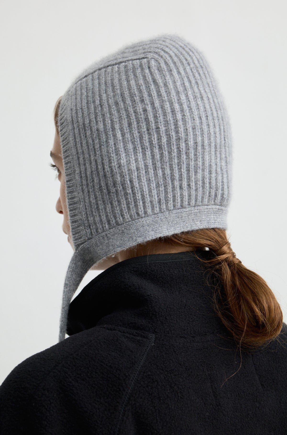 Woman wearing a grey knit hat with ties around the neck with a black flannel sweatshirt against a light grey wall