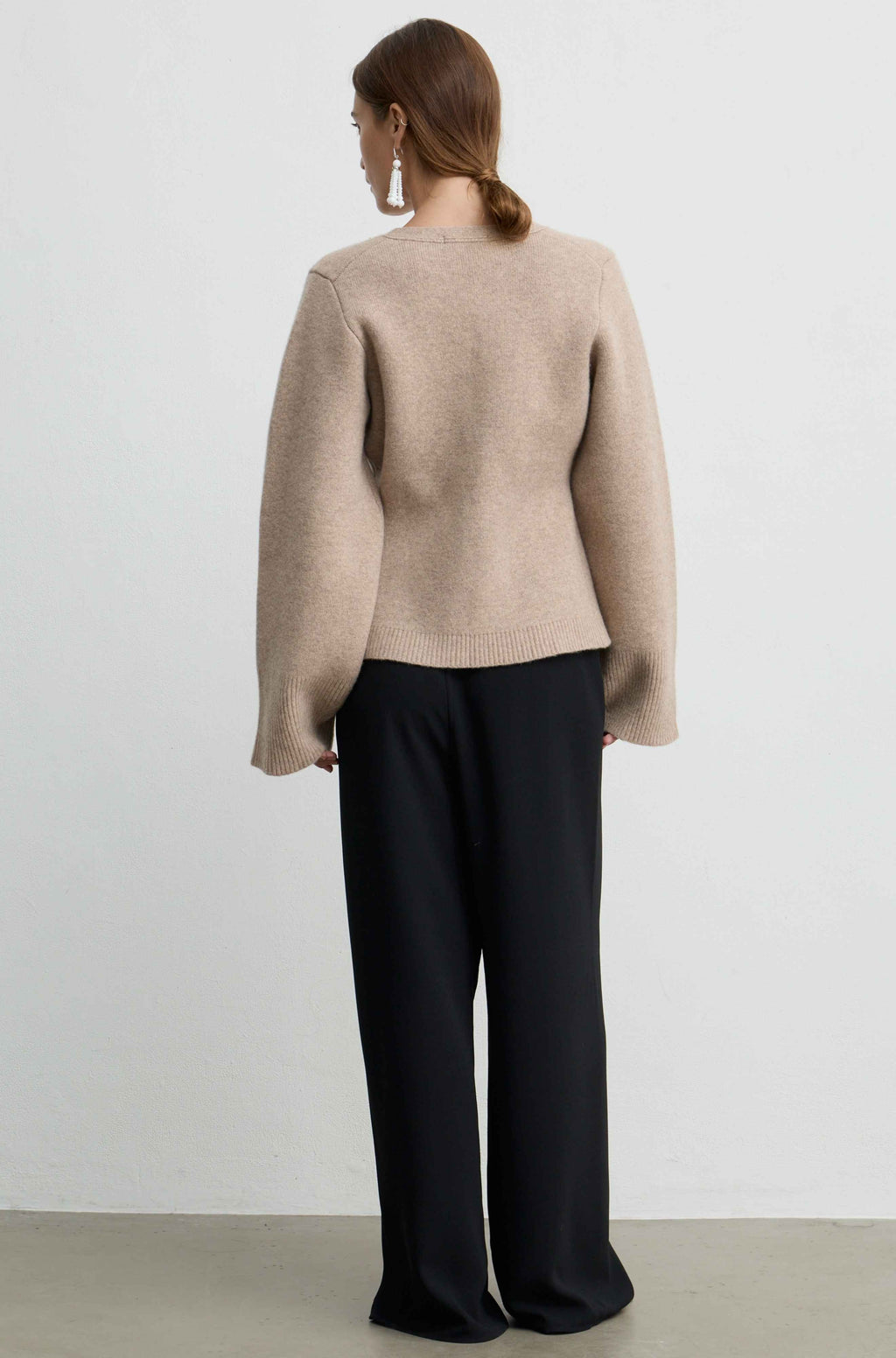 A woman shown from the back wearing a light beige knit sweater and black wide-leg trousers, standing against a plain light gray wall.
