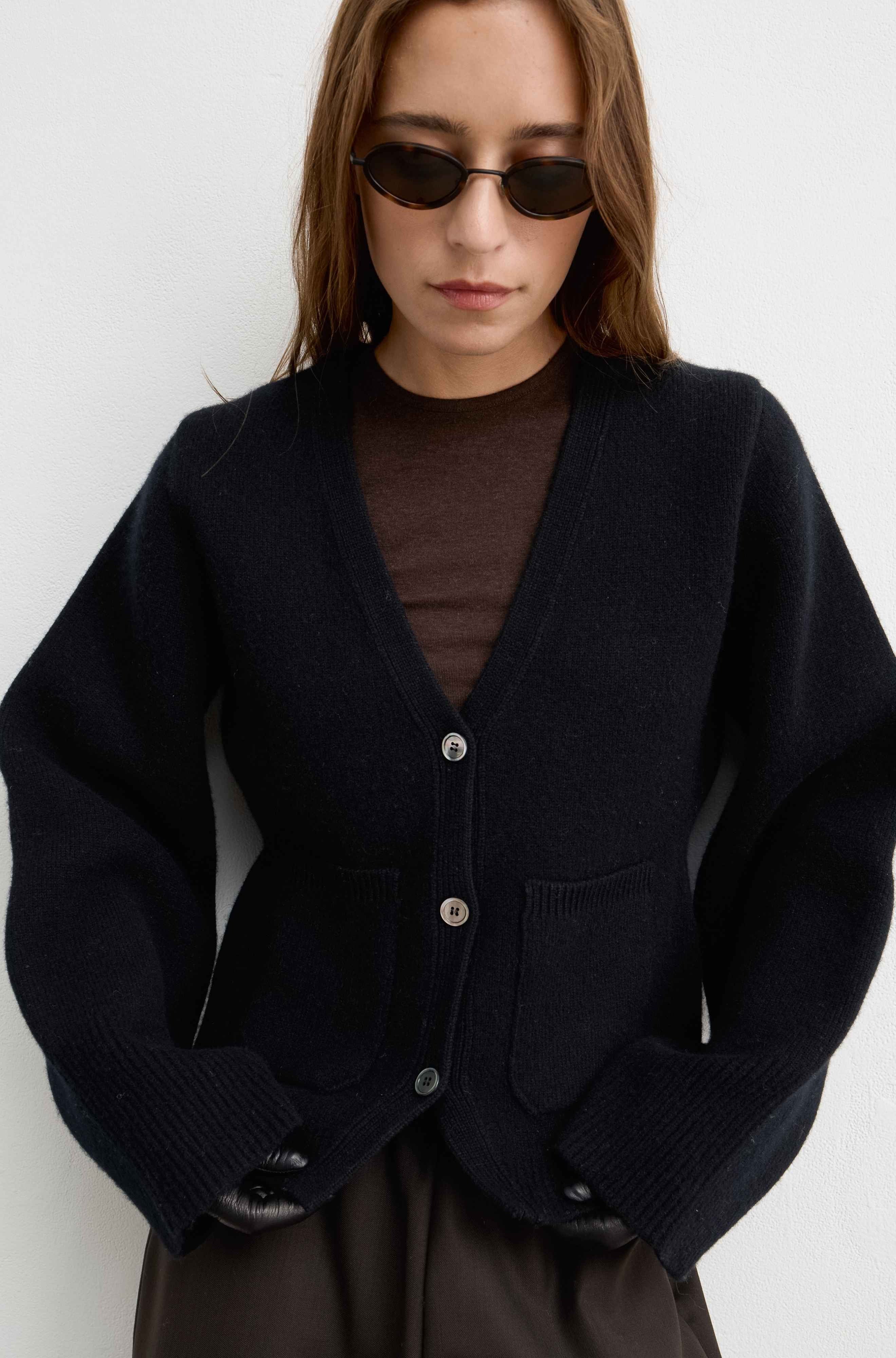 A woman wearing a black knit cardigan over a brown top, styled with dark sunglasses and a dark skirt, photographed against a plain light gray wall.