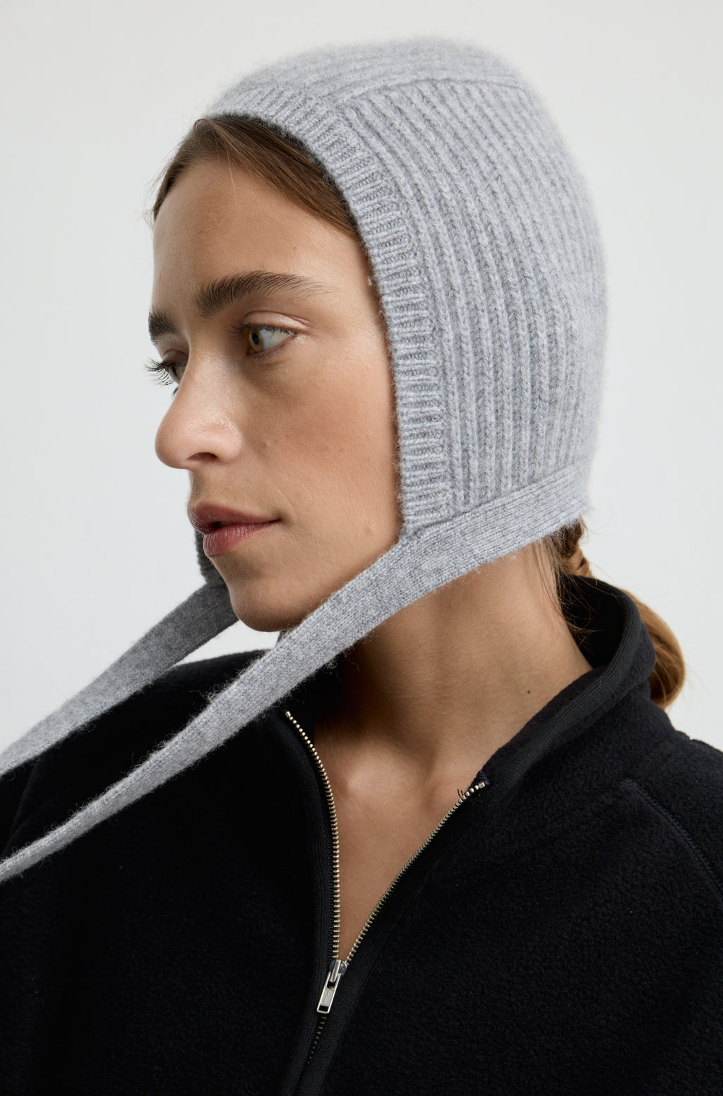 Woman wearing a grey knit hat with ties around the neck with a black flannel sweatshirt against a light grey wall