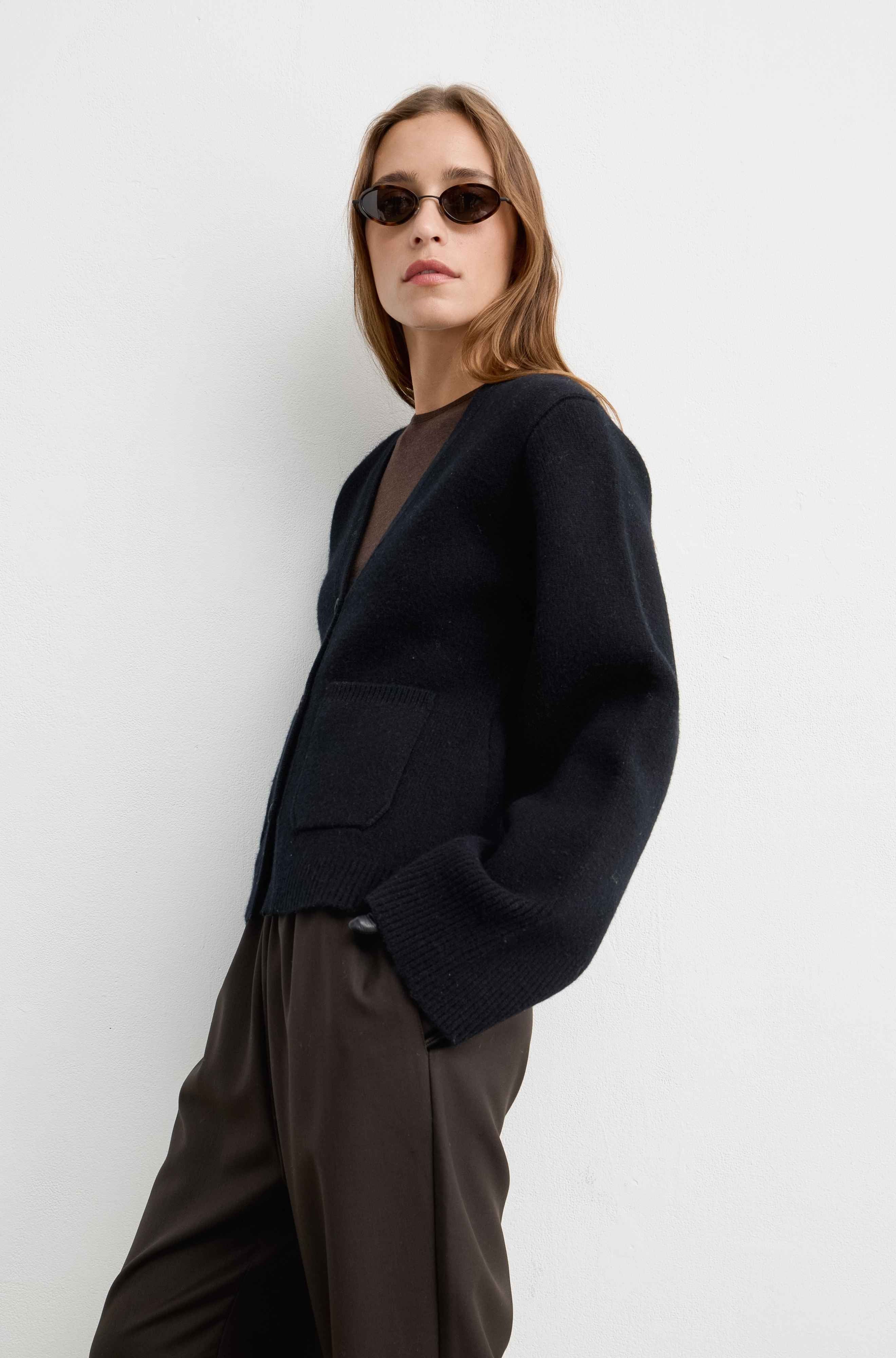 A woman modeling a black knit cardigan with front patch pockets, worn over a sheer brown top and paired with dark brown trousers, set against a plain light gray studio background.