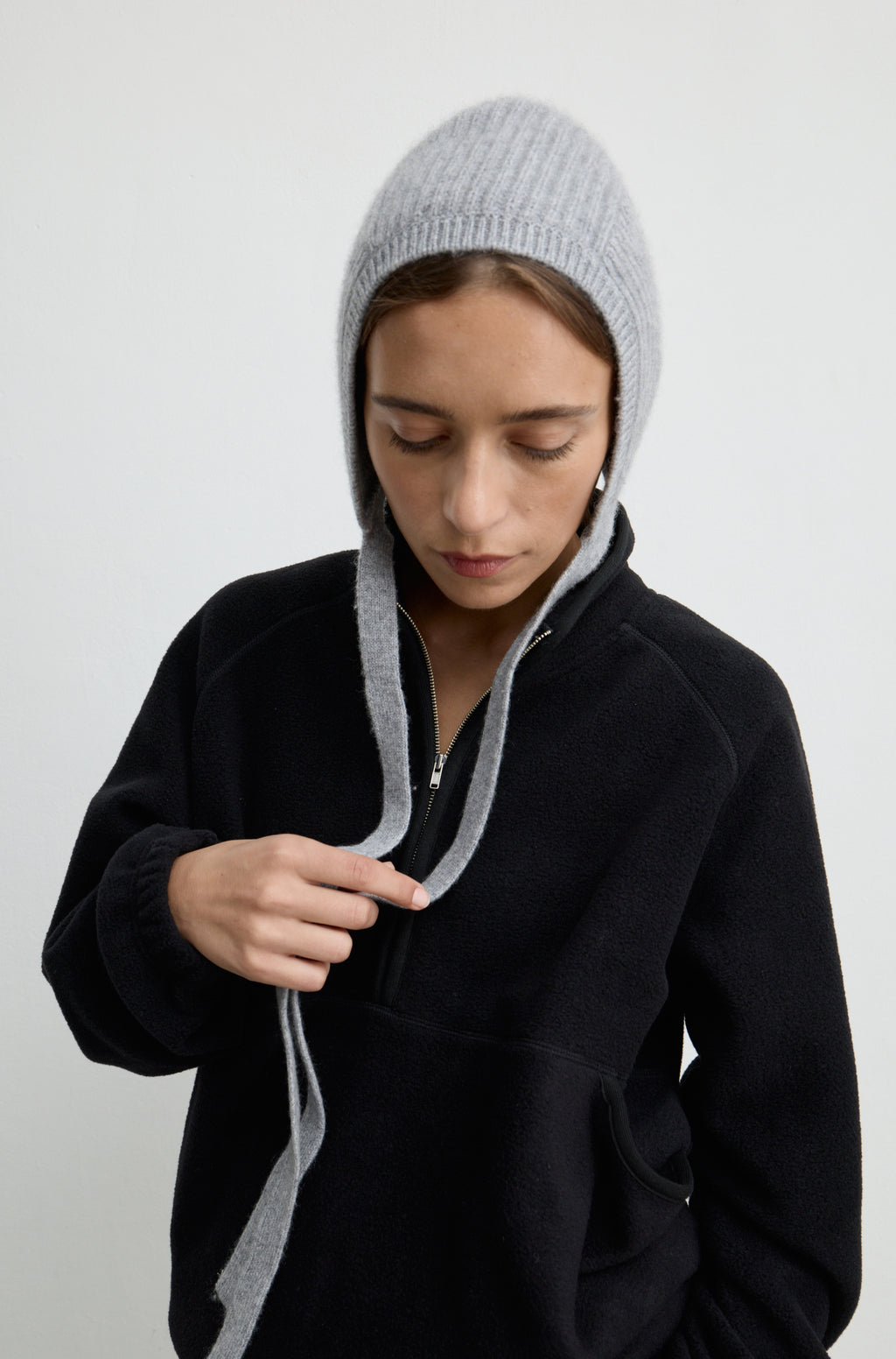 Woman wearing a grey knit hat with ties around the neck with a black flannel sweatshirt against a light grey wall