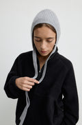 Woman wearing a grey knit hat with ties around the neck with a black flannel sweatshirt against a light grey wall
