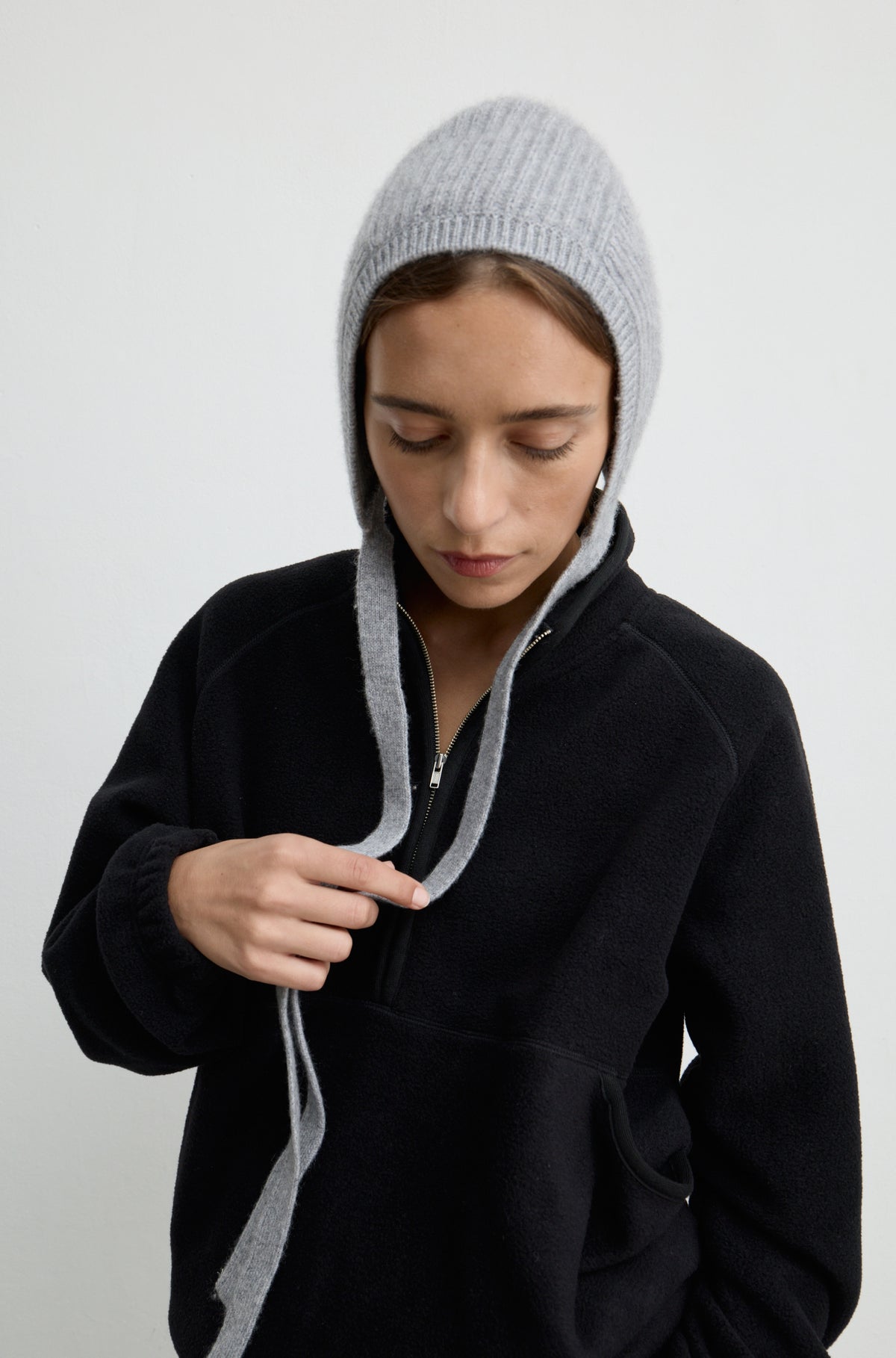 Woman wearing a grey knit hat with ties around the neck with a black flannel sweatshirt against a light grey wall