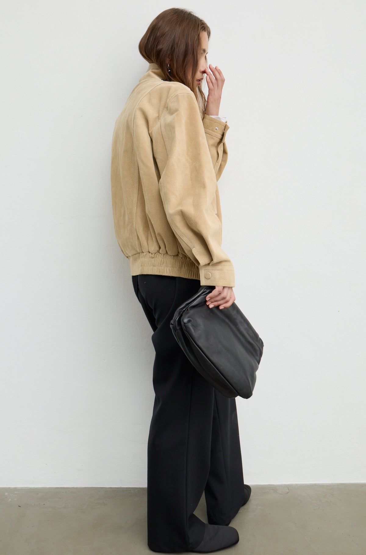 A woman stands in profile against a plain light wall, wearing a beige jacket, black wide-leg pants, and holding a black shoulder bag.