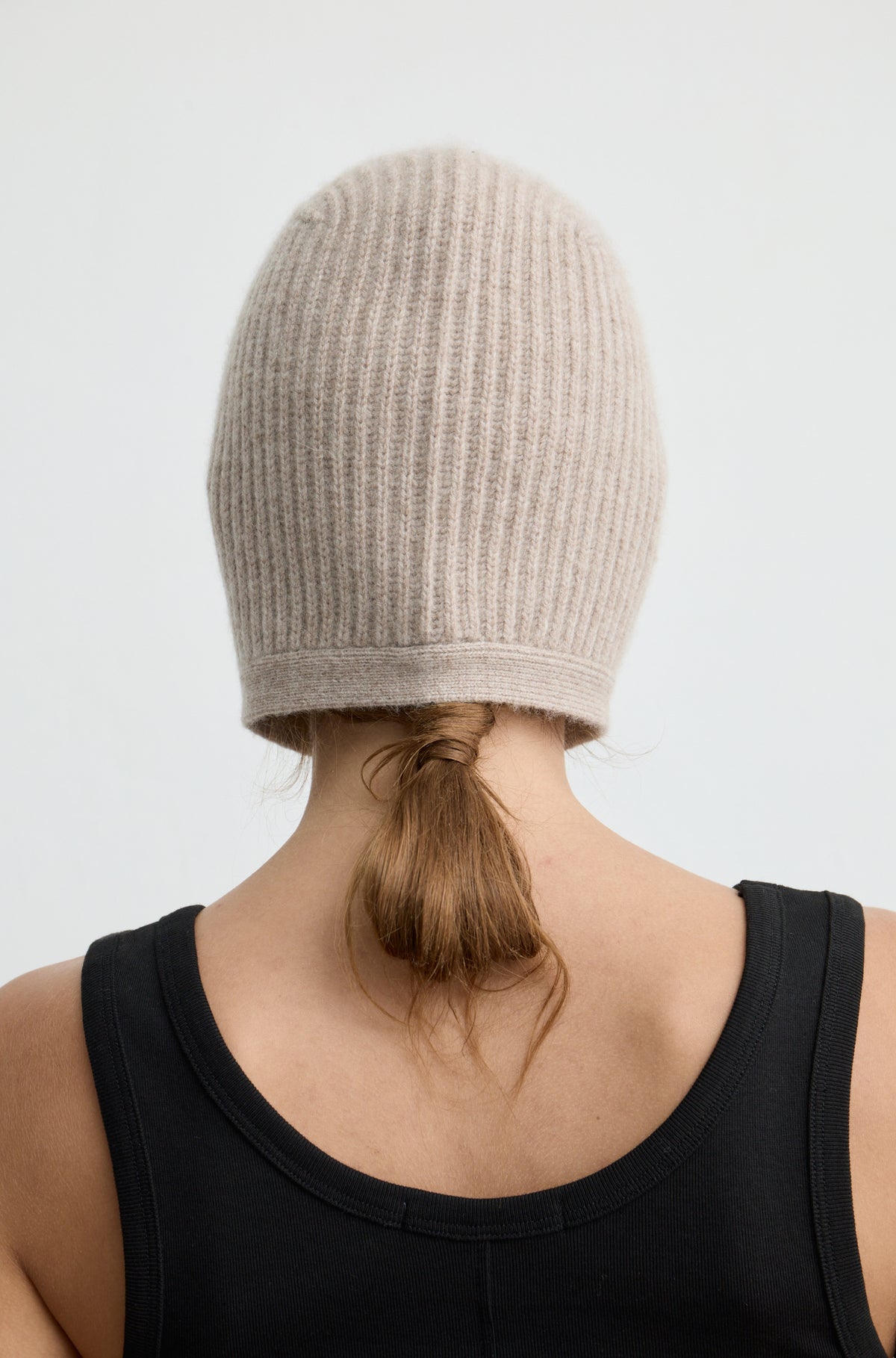 Woman wearing a light brown knit hat tied around the neck with a black tank top against a light grey wall
